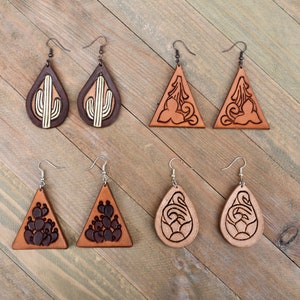 May include: Eight pairs of leather earrings with various designs, including cacti, flowers, and geometric patterns. The earrings are brown and tan in color and have silver hooks.