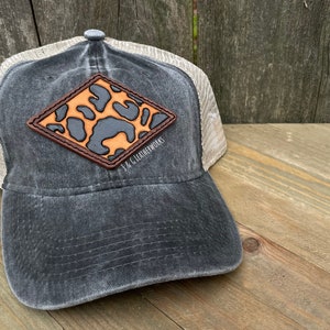 May include: A black and tan trucker hat with a diamond-shaped leather patch featuring a leopard print design. The patch has the text "J & G Leatherworks" in brown lettering.