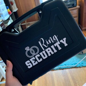 Könnte beinhalten: Schwarzer Hartschalenkoffer mit einem weißen Aufkleber, auf dem "Ring Security" steht, und einem stilisierten Bild von zwei ineinander verschlungenen Ringen mit einem Diamanten an einem Ring.