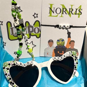 May include: White heart-shaped sunglasses with black lenses and green and black beads spelling out "LANDO NORRIS" on the frame. The sunglasses are on a blue surface.