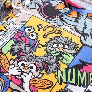 May include: A colorful t-shirt featuring a cartoon comic strip design with various characters and the word "NUMBER" in large green letters. The design includes yellow, blue, pink, and orange hues.