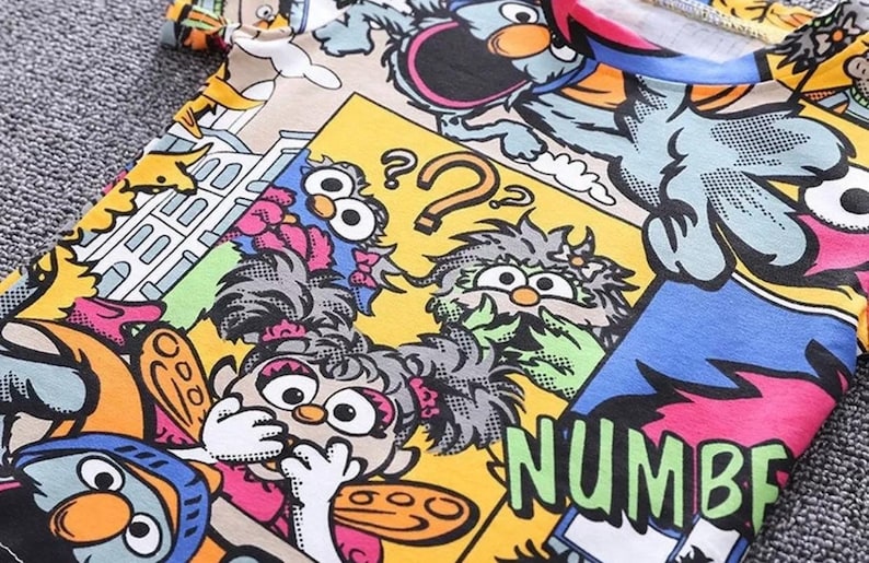 May include: A colorful t-shirt featuring a cartoon comic strip design with various characters and the word "NUMBER" in large green letters. The design includes yellow, blue, pink, and orange hues.