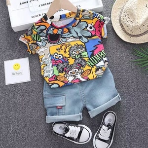 May include: A colorful children's outfit featuring a short-sleeved shirt with cartoon characters and denim shorts. The outfit includes black and white sneakers. A small card with a smiley face and the words "Be Happy" is also present.