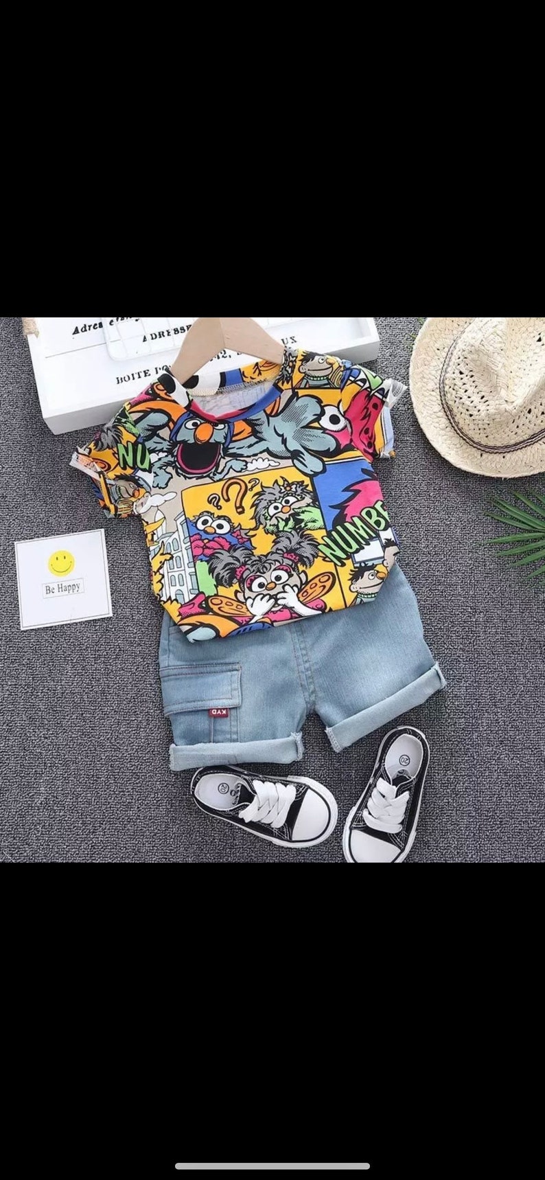 May include: A colorful children's outfit featuring a short-sleeved shirt with cartoon characters and denim shorts. The outfit includes black and white sneakers. A small card with a smiley face and the words "Be Happy" is also present.