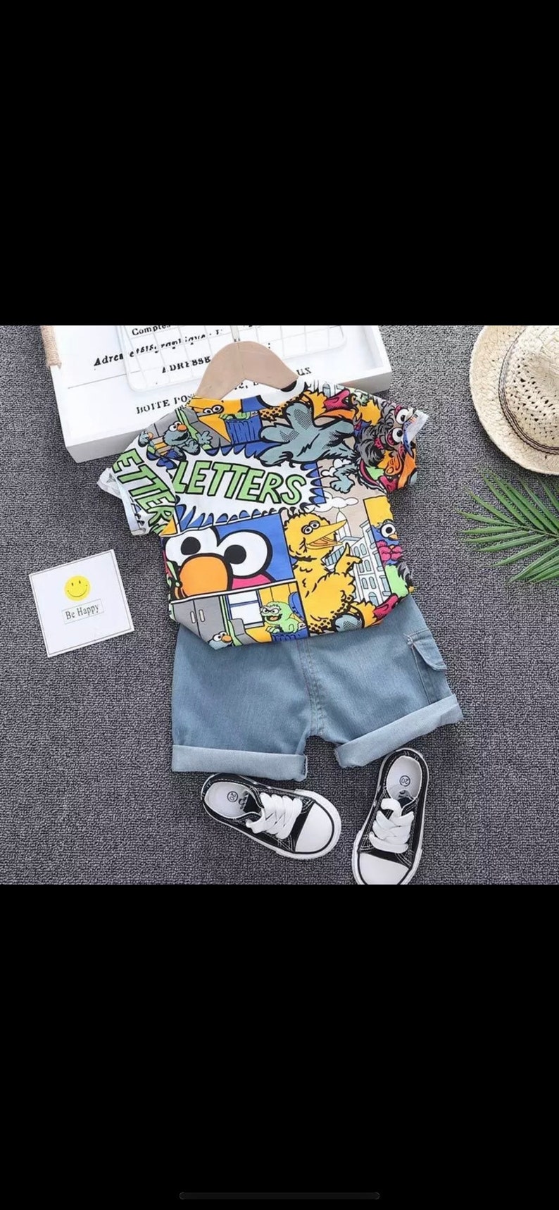 May include: A children's outfit featuring a colorful Sesame Street graphic t-shirt and denim shorts. The shirt has a vibrant print of characters and the word "LETTERS". Includes black and white sneakers.