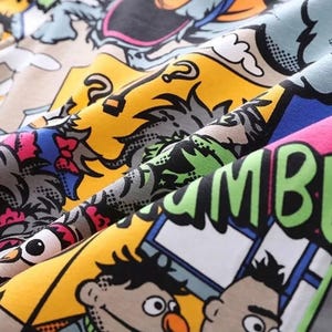 May include: Close-up of a colorful fabric with a comic book style print. The design features cartoon characters, speech bubbles, and bold lettering in various colors including yellow, blue, pink, and green. The fabric is folded, showing the pattern's details.