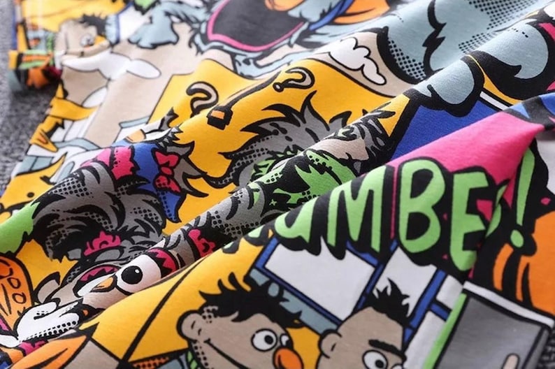May include: Close-up of a colorful fabric with a comic book style print. The design features cartoon characters, speech bubbles, and bold lettering in various colors including yellow, blue, pink, and green. The fabric is folded, showing the pattern's details.