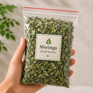 May include: A clear plastic resealable bag filled with dried green moringa leaves. A white square label on the front features a green leaf icon, the text "Moringa" in a bold dark green font, and the text "dried leaves" in a smaller font.