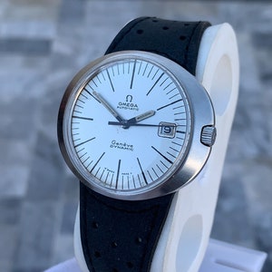May include: A silver Omega Genève Dynamic automatic wristwatch with a white face and black hands. The watch has a black leather band with a perforated pattern.