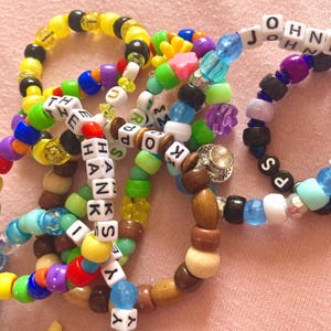 May include: A selection of colourful beaded bracelets. Some bracelets have lettered beads spelling out words and names. The beads are in various colours, including yellow, blue, green, and purple. The bracelets are made of different materials.