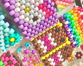 May include: A collection of colorful beaded bracelets with various patterns, including a purple and white striped bracelet, a brown and white checkered bracelet, and a rainbow striped bracelet.