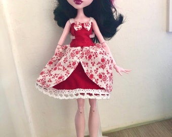 May include: A doll with black hair with pink highlights, wearing a red dress with a white floral pattern skirt and white lace trim. The doll has pale skin, dark eye makeup, and a heart-shaped mark on its cheek. It stands on a clear plastic stand.