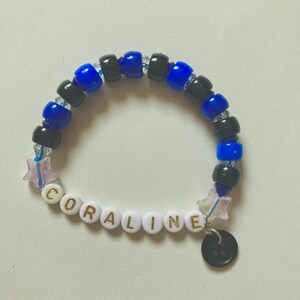 K&ouml;nnte beinhalten: Ein Perlenarmband mit dem Namen "CORALINE" in wei&szlig;en Buchstabenperlen. Das Armband hat blaue und schwarze Perlen, einen Sternanh&auml;nger und einen schwarzen Knopf. Ein handgefertigtes Accessoire.