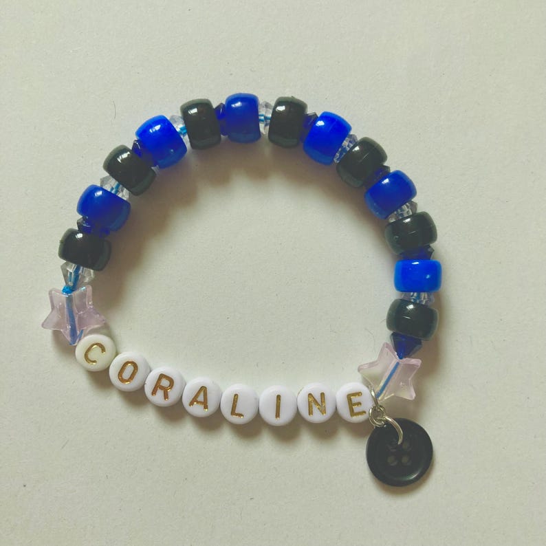 K&ouml;nnte beinhalten: Ein Perlenarmband mit dem Namen "CORALINE" in wei&szlig;en Buchstabenperlen. Das Armband hat blaue und schwarze Perlen, einen Sternanh&auml;nger und einen schwarzen Knopf. Ein handgefertigtes Accessoire.