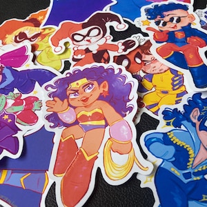 May include: A selection of vibrant cartoon stickers depicting superheroes and comic book characters. The stickers feature characters such as Wonder Woman, Harley Quinn, and Superman, with bright colours and detailed illustrations. The stickers are on a black surface.