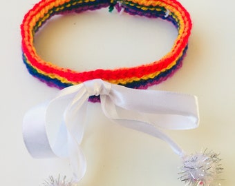 Può includere: Un collare all'uncinetto arcobaleno con un nastro di raso bianco e due pompon argentati.