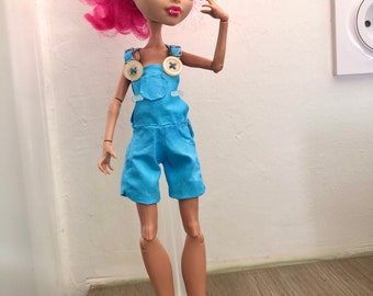 May include: A fashion doll with pink hair wearing a light blue overall outfit with two large buttons. The doll has brown skin and is standing on a clear plastic stand.