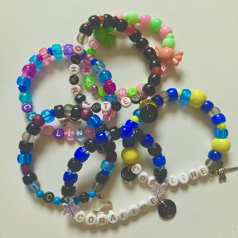K&ouml;nnte beinhalten: Eine Sammlung farbenfroher Perlenarmb&auml;nder. Die Armb&auml;nder weisen verschiedene Farben auf, darunter Blau, Rosa, Schwarz und Gelb, wobei einige Namen wie "CORALINE" und "LINE" buchstabieren. Charms und dekorative Perlen sorgen f&uuml;r Details.