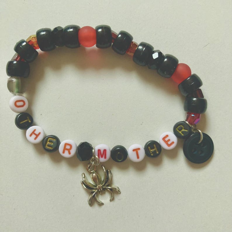 K&ouml;nnte beinhalten: Ein Armband mit schwarzen, roten und wei&szlig;en Perlen. Das Armband buchstabiert "TO HER MOTHER" mit Buchstabenperlen. Ein silberner Spinnenanh&auml;nger und ein schwarzer runder Anh&auml;nger h&auml;ngen an dem Armband.