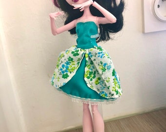 May include: A doll with pink and black hair wearing a teal dress with a white floral pattern. The doll has a heart drawn on her cheek.