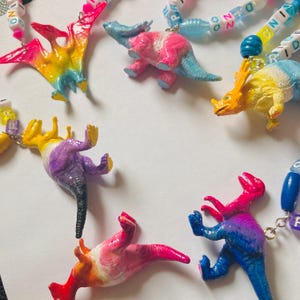 Pode incluir: Uma coleção de pingentes em forma de dinossauro presos a colares de contas. Cada dinossauro de plástico é pintado em gradientes de cores vibrantes. As contas incluem blocos quadrados brancos com letras pretas soletrando PRIDE e DINO.