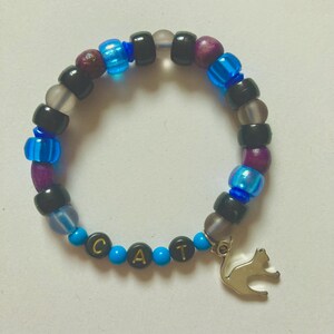 K&ouml;nnte beinhalten: Ein Perlenarmband mit einem silbernen Katzenanh&auml;nger. Das Armband hat eine Mischung aus schwarzen, blauen, lila und transparenten Perlen. Das Wort "CAT" ist mit goldenen Buchstaben auf blauen Perlen geschrieben.
