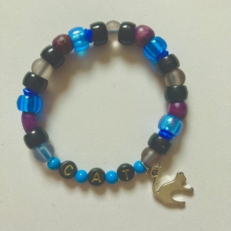 K&ouml;nnte beinhalten: Ein Perlenarmband mit einem silbernen Katzenanh&auml;nger. Das Armband hat eine Mischung aus schwarzen, blauen, lila und transparenten Perlen. Das Wort "CAT" ist mit goldenen Buchstaben auf blauen Perlen geschrieben.