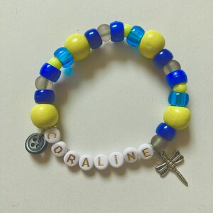 K&ouml;nnte beinhalten: Ein Armband mit gelben, blauen und transparenten Perlen. Das Armband tr&auml;gt den Namen "CORALINE" in wei&szlig;en Buchstabenperlen. Ein silberner Libellen-Anh&auml;nger und ein Knopf-Anh&auml;nger sind angebracht.