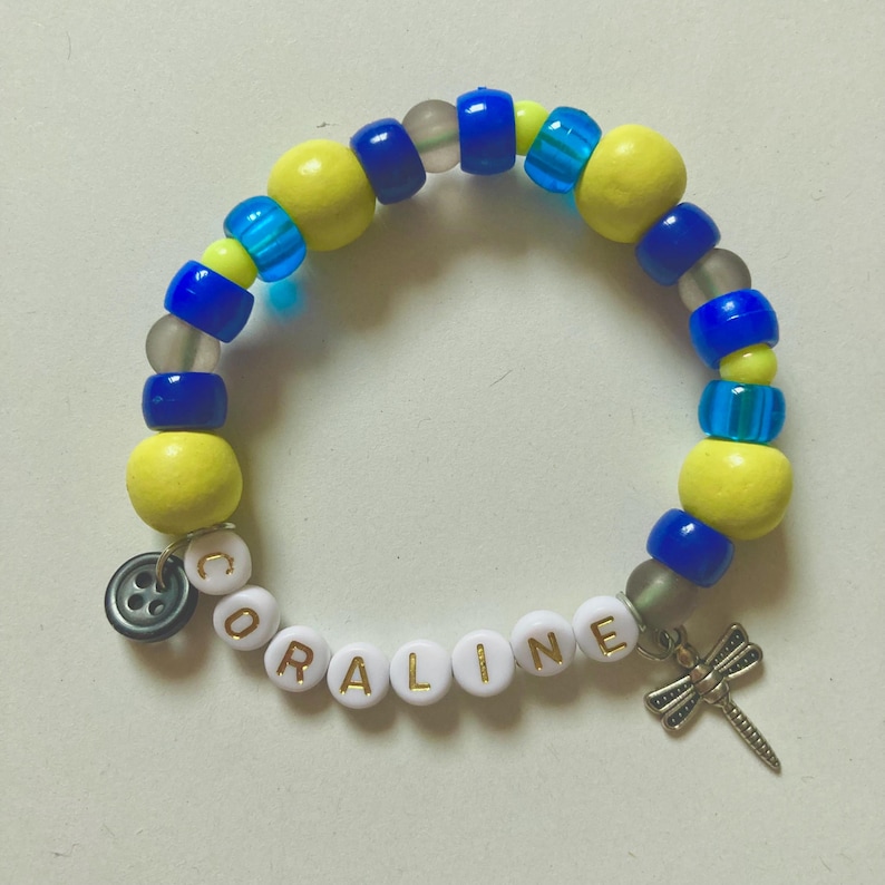 K&ouml;nnte beinhalten: Ein Armband mit gelben, blauen und transparenten Perlen. Das Armband tr&auml;gt den Namen "CORALINE" in wei&szlig;en Buchstabenperlen. Ein silberner Libellen-Anh&auml;nger und ein Knopf-Anh&auml;nger sind angebracht.