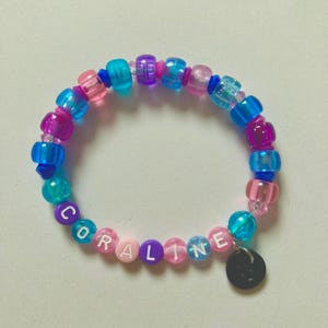 K&ouml;nnte beinhalten: Ein farbenfrohes Perlenarmband mit dem Namen "CORALINE" in wei&szlig;en Buchstabenperlen. Das Armband besteht aus rosa, blauen und lila Perlen und hat einen kleinen schwarzen Anh&auml;nger. Ein fr&ouml;hliches und farbenfrohes Accessoire.