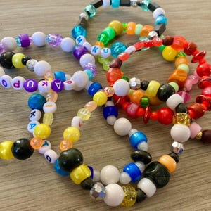 May include: A close-up of several colourful beaded bracelets with different letter beads spelling out names. The bracelets are made with white, yellow, blue, green, red, orange, and purple beads.