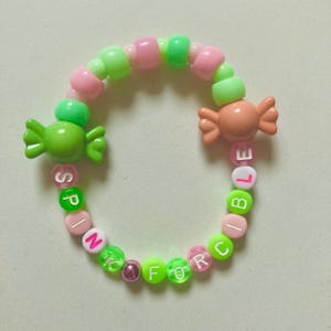 K&ouml;nnte beinhalten: Ein farbenfrohes Perlenarmband mit rosa, gr&uuml;nen und wei&szlig;en Perlen. Das Armband hat bonbonf&ouml;rmige Charms und Buchstabenperlen mit der Aufschrift "SPINK FORCE BLE". Ein verspieltes und farbenfrohes Accessoire.