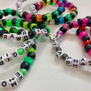 Può includere: Cinque braccialetti di perline colorate con perline di lettere bianche che compongono le parole "RAW", "NOT A PHASE", "DOWN", "GIRL" e "EMO".