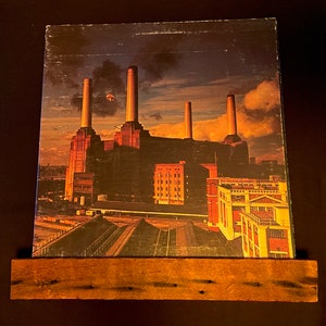 May include: A vintage vinyl record album cover featuring a photograph of a brick building with tall smokestacks. The building is a power plant, and the album cover is titled "Animals" by Pink Floyd.