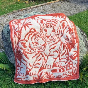 Puede incluir: Una manta cuadrada de ganchillo con un diseño de dos tigres en tonos naranja y blanco. Los tigres están rodeados de bambú y otro follaje. La manta tiene un borde naranja liso.