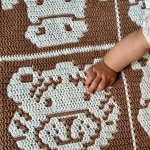 K&ouml;nnte beinhalten: Eine braun-wei&szlig;e H&auml;keldecke mit einem sich wiederholenden Muster eines Kuhgesichts. Die Decke ist mit einem strukturierten Stich gefertigt und hat ein weiches Aussehen. Die Hand eines Babys ber&uuml;hrt die Decke.