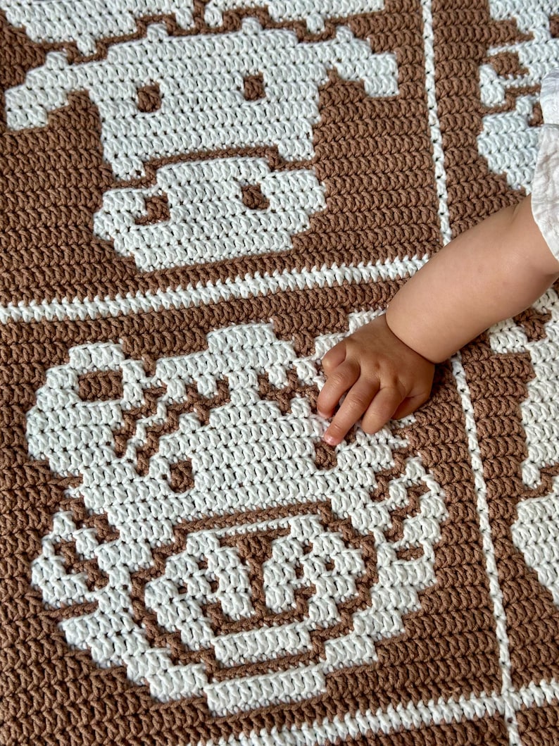 K&ouml;nnte beinhalten: Eine braun-wei&szlig;e H&auml;keldecke mit einem sich wiederholenden Muster eines Kuhgesichts. Die Decke ist mit einem strukturierten Stich gefertigt und hat ein weiches Aussehen. Die Hand eines Babys ber&uuml;hrt die Decke.