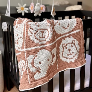 K&ouml;nnte beinhalten: Eine handgefertigte Babydecke mit Tiergesichtern in einem Kinderbett. Die Decke hat ein sich wiederholendes Muster aus wei&szlig;en Tiergesichtern auf hellbraunem Hintergrund. Das Kinderbett ist schwarz, und ein Mobile mit Blumen und einem Hasen h&auml;ngt dar&uuml;ber.