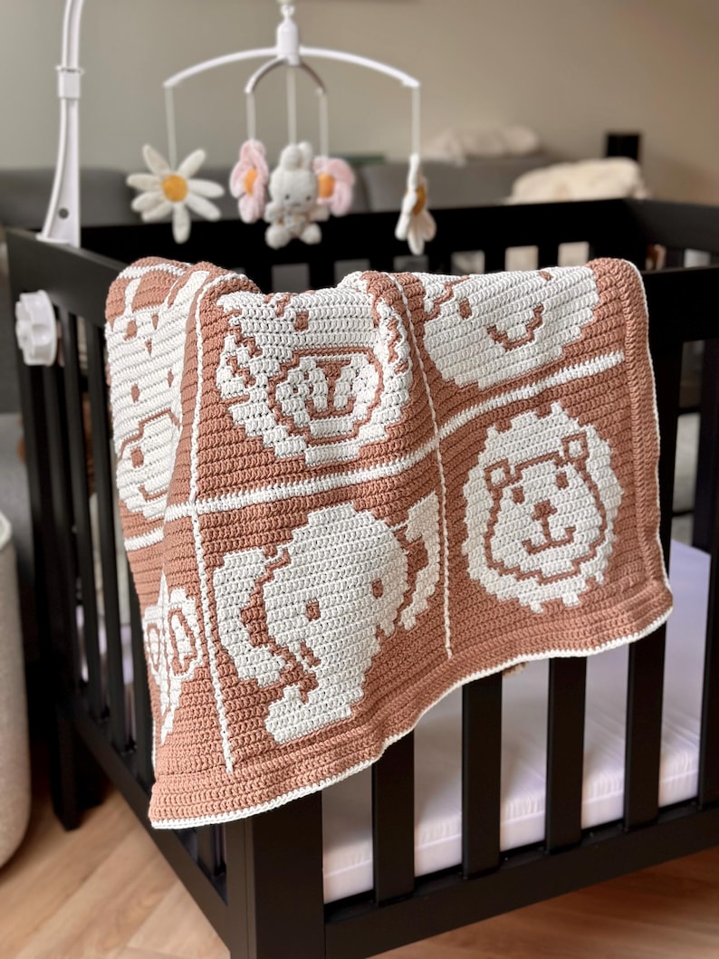 K&ouml;nnte beinhalten: Eine handgefertigte Babydecke mit Tiergesichtern in einem Kinderbett. Die Decke hat ein sich wiederholendes Muster aus wei&szlig;en Tiergesichtern auf hellbraunem Hintergrund. Das Kinderbett ist schwarz, und ein Mobile mit Blumen und einem Hasen h&auml;ngt dar&uuml;ber.