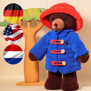 Könnte beinhalten: Ein handgefertigter brauner Teddybär, der einen blauen Mantel mit roten Knebeln und einen roten Hut trägt. Der Bär hat eine hellbraune Schnauze und steht vor einem Holzausschnitt eines Baumes. Der Bär ist ein Plüschtier.