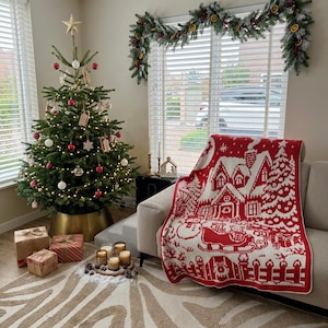 May include: A Christmas tree decorated with red and white ornaments, a gold star topper, and warm lights, standing in a gold metal base. A red and white knit throw blanket with a winter village scene is draped over a sofa next to wrapped gifts.