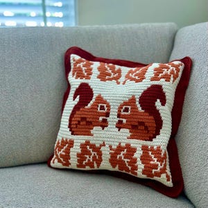 Puede incluir: Un cojín cuadrado de ganchillo color crema con un borde rojo. El cojín presenta dos ardillas enfrentadas en tonos naranja y marrón, con patrones de hojas arriba y abajo. El cojín está sobre un sofá gris.