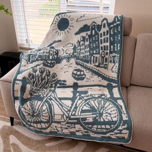 May include: A rectangular crochet blanket featuring a two-tone teal and cream design. The pattern depicts a city canal scene with tall buildings, a sun, a bridge, and a bicycle in the foreground with a basket filled with flowers on the handlebars.