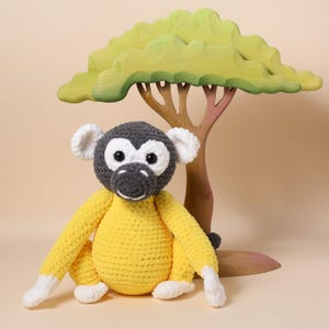 May include: A handmade crochet monkey with a yellow body, gray head, and white accents. The monkey sits in front of a wooden tree with a green canopy. The toy is soft and plush, perfect for children.