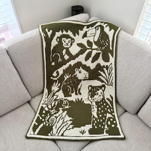 Könnte beinhalten: Eine gehäkelte Decke mit einem Dschungelmotiv, das einen Affen, einen Tukan und andere weiße Tiere auf einem dunkelgrünen Hintergrund zeigt. Die Decke hat einen weißen Rand und liegt auf einem hellgrauen Sofa.