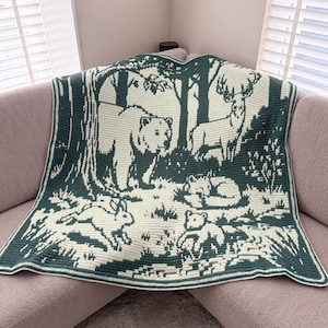 May include: A forest-themed crochet blanket in shades of green and cream. The blanket features a detailed design of bears, deer, rabbits, and trees. The animals are depicted in a woodland scene, creating a cozy and nature-inspired aesthetic.