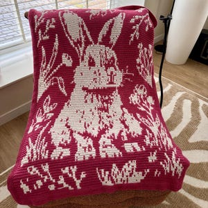 May include: A burgundy and white crochet blanket draped over a chair. The blanket features a large white rabbit design surrounded by floral and plant motifs. The blanket is made of soft yarn and is suitable for home decor.
