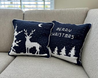May include: Two navy blue crochet pillows with white accents. One pillow features a deer silhouette under a crescent moon and stars. The other pillow says "MERRY CHRISTMAS" above a row of trees. Both pillows have white borders.