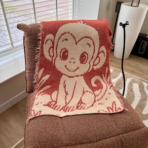 May include: A red and cream-colored knitted throw blanket featuring a smiling monkey design. The blanket is draped over a brown chair, with the monkey's face and body prominently displayed. The blanket has a fringed edge.