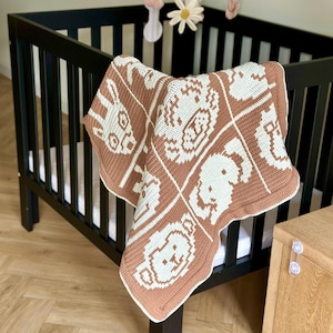 K&ouml;nnte beinhalten: Eine handgefertigte Babydecke aus H&auml;kelarbeit in Braun und Wei&szlig; mit Tiermustern. Die Decke liegt &uuml;ber der Seite eines schwarzen Kinderbetts. &Uuml;ber dem Bett h&auml;ngt ein Mobile mit Blumen- und Sternformen.